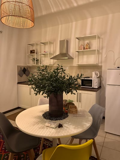The image shows an interior scene of a kitchen. The style is modern with white cabinets, shelving, and appliances. A round table is the focal point, surrounded by chairs in grey and yellow. On the table sits a large vase with greenery. Above the table is a rattan pendant light. The kitchen appears to be well-lit. While the precise location within Perugia, Italy, cannot be determined from the image, the interior design suggests a contemporary apartment or house. No people are visible in the image.