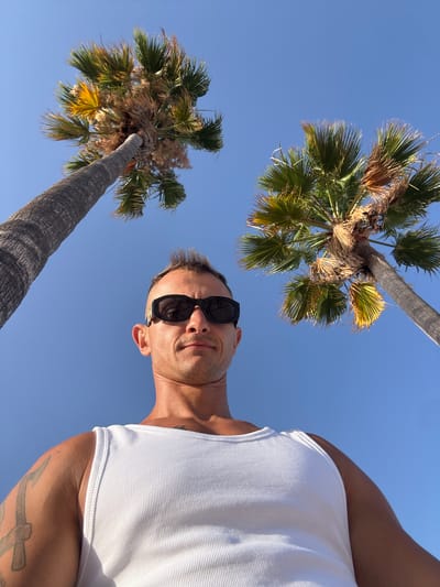 This is a low-angle, upside-down selfie of a man wearing sunglasses and a white tank top. He is outdoors with a clear blue sky and palm trees in the background, suggesting a warm, sunny day. The man appears to be in his 30s or 40s, with short, styled hair and stubble. He is looking towards the camera with a slight smile. Visible on his left arm is a tattoo of an anchor with a cross. The overall impression is one of a relaxed, vacation-like atmosphere, possibly in a coastal location. The presence of palm trees and the clear blue sky strongly indicate a Mediterranean setting, aligning with the provided location context of Altea, Spain. The lighting suggests it is daytime, likely late morning or afternoon due to the brightness and shadows.
