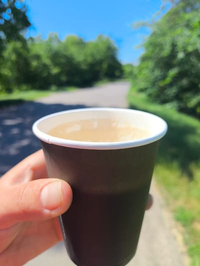A close-up shot captures a person's hand holding a dark brown paper cup filled with a light-colored beverage, possibly coffee or tea. The cup has a white rim. The background is blurred, revealing a sunlit outdoor scene. A paved pathway winds through lush green trees under a bright blue sky. The sun casts dappled shadows on the path, indicating a daytime setting. The foliage suggests a park, nature trail, or a similar recreational area. The overall mood is relaxed and serene, hinting at a moment of quiet enjoyment outdoors, perhaps during a walk. There is no visible text in the image. The location context is Anastasivka, Ukraine.