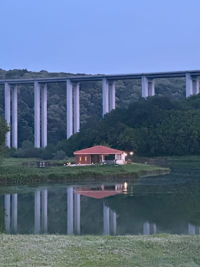 A tranquil twilight scene in Pisanets, Bulgaria, features a multi-pier concrete viaduct bridge spanning a densely forested valley under a clear, light blue sky. In the midground, a small, single-story building with a red-tiled roof and illuminated wall sconces rests on a grassy peninsula extending into a calm body of water. The water reflects the bridge's vertical piers and the building's warm lights. A small, dark platform structure is visible on the water's edge to the left of the building. The immediate foreground consists of a strip of mown green grass. No people or animals are visible.