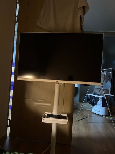 An indoor scene, possibly a residential living space or storage area in Nonsan-si, South Korea, is depicted. A large, black television screen is mounted on a white, floor-standing television stand. A remote control rests on a small shelf attached to the stand. To the right of the television stand, a clothes drying rack holds various garments, and a white appliance, possibly a dehumidifier or air purifier, is visible on the floor. A wooden door is open to the left of the television, revealing a sliver of a brightly lit hallway or adjacent room. A beige towel is draped over the top of the door. The flooring appears to be wood laminate.