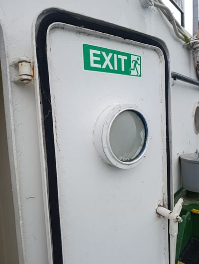 A white metal door is visible on a ship or industrial structure. The door has a round porthole window with a blue trim, and an "EXIT" sign with a running person icon is affixed to it. A black rubber seal runs along the edge of the doorframe. A white latch mechanism is attached to the right side of the door. The background shows a white wall and some pipes. The scene appears to be outdoors, likely in an industrial or marine environment. There is no indication of this scene being located in the city of Šabac, Serbia.