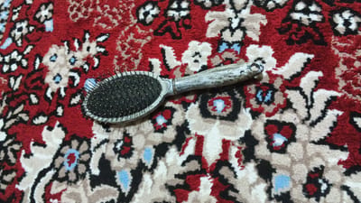 A hairbrush with a patterned, silver-toned handle lies diagonally on a red, cream, and black patterned carpet. The bristles of the hairbrush are black and metal. The carpet has a floral or paisley design. There are no people visible in the image, and no context for a specific location in Abdurahmoni Jomi, Tajikistan, is provided by the image itself.