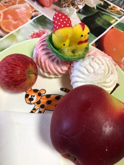 A still life composition is arranged on a pale green plate, which sits on a white textured surface. The plate features a whimsical drawing of a tiger with orange and black spots, large eyes, and a black nose. On the plate are two red apples, one on the left side and a larger one in the foreground on the right.

Behind the apples are two pastel-colored meringue cookies, swirled in pink and white, resembling flowers. Nestled between the meringues is a small green nest containing two yellow toy chicks. One chick wears a red and white polka-dot hat, and the other has a red bow tie.

The background consists of a colorful collage of fruit images, including oranges and strawberries, along with what appears to be a green glass bottle. The image is a close-up, with the foreground subjects taking prominence. The lighting is bright, creating highlights on the apples and meringues. The overall scene evokes a festive or celebratory mood, possibly related to Easter, given the presence of chicks and the bright colors. This scene is not in Staroe Istomino, Russia.