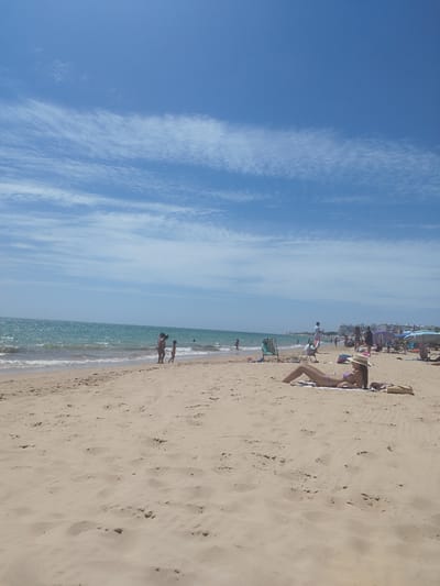 The image depicts a sunny day at a crowded beach in Rota, Spain. The foreground is dominated by sand, with footprints visible. In the midground, several people are scattered across the beach and in the ocean. A woman in a purple bikini and a straw hat is sunbathing on a towel. Further back, a family, possibly a mother and two children, are near the water's edge, with one child appearing to be walking towards the ocean. The ocean itself is a vibrant blue-green, with moderate waves. In the background, several beach umbrellas and tents are set up, and buildings line the distant shore. The sky is a clear blue with wispy white clouds.
