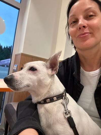 An indoor selfie shows a woman and a white Jack Russell Terrier. The woman, with dark hair and a black denim jacket over a white ribbed tank top, is smiling and looking at the camera. The dog, looking to the left, wears a black collar with silver studs and a leash clip. The background includes a window with a view of trees and a light blue sky, and a hint of a sign with Cyrillic text that may indicate the location. The setting appears to be a cafe or restaurant with wooden accents. The image was taken in Chusovoy, Russia.