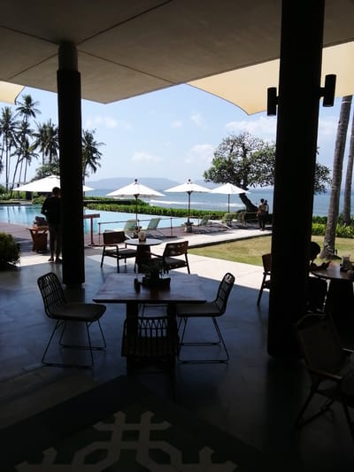 The image depicts an outdoor seating area at what appears to be a resort or restaurant overlooking a swimming pool and the ocean. The foreground features several tables and chairs, some made of dark metal and rattan, positioned on a tiled floor. Beyond this seating area, a swimming pool with lounge chairs and umbrellas is visible, adjacent to a lush green lawn and palm trees. The ocean stretches to the horizon under a clear blue sky with scattered clouds. In the background, a person stands near the pool, facing away from the camera. The architecture suggests a tropical setting, consistent with locations in Banyuwangi, Indonesia, known for its coastal resorts. The lighting indicates it is daytime.
