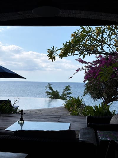 This is a photograph of an outdoor seating area with a view of the ocean in Gerokgak, Indonesia. In the foreground, there is a dark, silhouetted lounge area with wicker furniture and a glass-topped coffee table. On the coffee table sits a dark, metallic hookah. Beyond the seating area, a wooden deck leads to an infinity pool that appears to merge with the sea. Lush green and purple flowering plants frame the view of the calm, blue ocean under a bright, cloudy sky. A large black umbrella is visible to the left of the pool. The overall scene suggests a relaxed, tropical atmosphere, likely a resort or private villa.