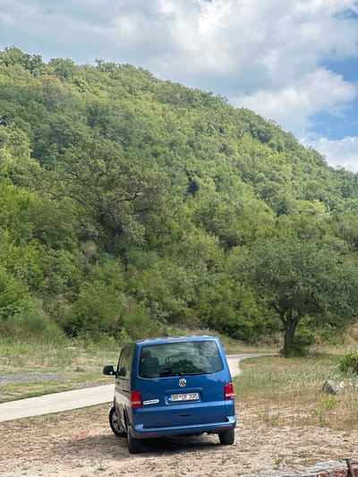 A blue Volkswagen van is parked on a gravelly surface next to a paved pathway. The van faces away from the viewer, showing its rear with a license plate reading "BR DF 395". The van has two stickers on its rear bumper. To the left of the van, a road curves away, disappearing into the distance. The van is positioned in front of a dense, green forest that covers a steep hillside. The sky above is partly cloudy with patches of blue. The surrounding vegetation includes various shades of green trees and bushes. The area appears to be a rural or natural setting, possibly a park or a scenic overlook, located in Čučuci, Montenegro.