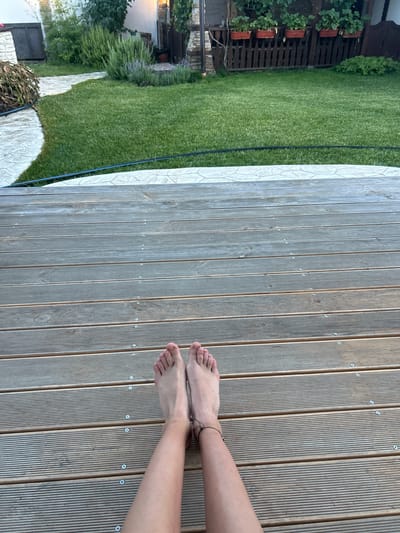 A pair of bare feet rests on a wooden deck. The feet are positioned close together, with the toes pointing upwards. The deck is made of wooden planks with visible screws. Beyond the deck is a green lawn with a stone path curving to the left. In the background, there is a fence with potted plants and some greenery. The scene appears to be a residential backyard. The location is Войводино, Bulgaria.
