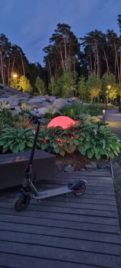In Barvikha, Russia, at dusk, an electric scooter is parked on a wooden deck in front of a garden. The scooter is dark gray and black with red accents. Behind the scooter, large green plants surround a glowing red sphere, possibly a decorative light. In the background, a rocky slope is illuminated by yellow lights, and tall, slender pine trees line the top of the slope. The sky is a deep blue, indicating twilight.