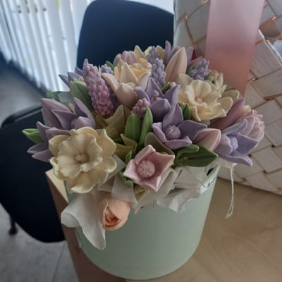 A close-up, high-angle shot captures a meticulously crafted bouquet of artificial flowers arranged in a pale green cylindrical container. The flowers are made of a delicate material, possibly fondant or frosting, and come in a pastel palette of soft yellows, lavenders, pale pinks, and light peaches. Among the blossoms are textured clusters resembling hyacinths, and petal designs that mimic tulips and daffodils. Green fondant leaves are interspersed throughout the arrangement, adding a touch of natural contrast.

The bouquet is partially cradled within a woven basket, its texture and color suggesting it might be made of straw or a similar natural material. A pale pink ribbon is draped across the basket, adding a soft decorative element. A small white ribbon with faint text, likely a brand or "handmade" label, is visible attached to the bouquet's wrapping.

The setting appears to be indoors, with the bouquet resting on a wooden surface, possibly a table. In the background, a dark chair is visible, and to the left, a white slatted structure, reminiscent of blinds, suggests a window. The overall lighting is soft and diffused, indicating it might be daytime, though no specific time can be determined. The composition evokes a sense of gentle beauty and artisanal skill. No people are visible in the image.