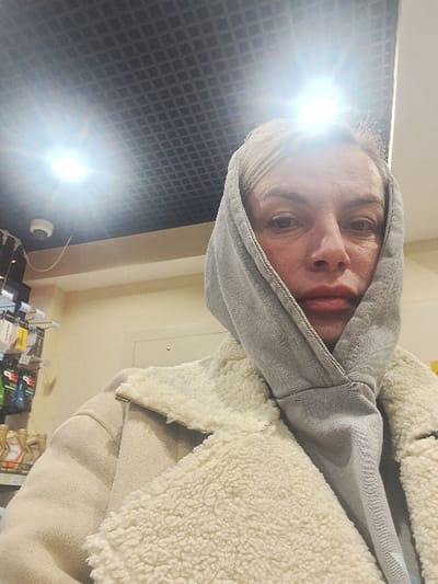 A person in a gray hoodie with the hood up and a sherpa-lined jacket. The individual's face is visible with pale skin and neutral expression. There's a bright glare in the upper left corner, possibly from a light source. The background features a dark, textured wall on the left and beige-colored walls and what appears to be a shelf or counter with items on the right.