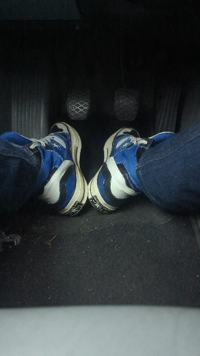 An overhead view captures a person's feet and lower legs inside a car's driver footwell. The individual wears blue denim jeans and distinctive high-top sneakers that are primarily blue, white, and black. A white star logo is visible on the side of both shoes, and a black patch with a white star is on the back heel of the soles.

The feet are positioned near two car pedals, which have a diamond or cross-hatch textured pattern. The floor of the car appears to be dark, likely black carpet. The scene suggests the person is either driving or preparing to drive. The lighting is dim, characteristic of a vehicle's interior. No specific location cues for Sélestat, France are discernible within the image. There is no clearly legible text beyond the star logos on the sneakers.