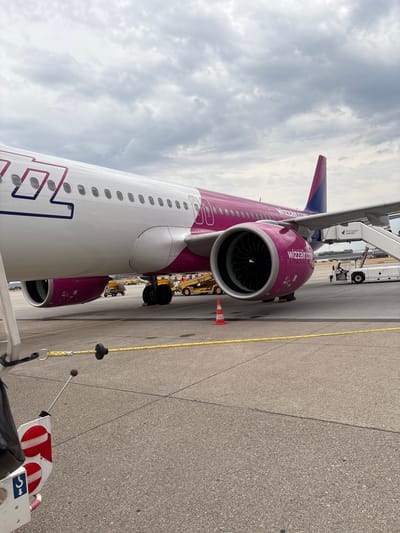 This is a photograph taken at Stuttgart Airport in Echterdingen, Germany, on a cloudy day. The primary subject is the fuselage and engine of a Wizz Air airplane, predominantly colored white and pink. The aircraft is parked on the tarmac, with the registration "wizzair.com" visible on the engine nacelle.

In the background, service vehicles are present, including a baggage tug and a mobile stairs leading to the aircraft's door. The sign on the mobile stairs indicates "Stuttgart Airport." There are no people clearly visible in the immediate vicinity of the plane's entrance.

In the foreground, a yellow rope barrier separates the viewing area from the aircraft. A portion of what appears to be ground equipment, possibly part of a chocking system or a pushback tractor attachment, is visible on the left side of the frame, displaying red and white striped safety markings. The tarmac is paved and shows clear demarcation lines. The overall impression is one of an operational airport environment under a slightly overcast sky.