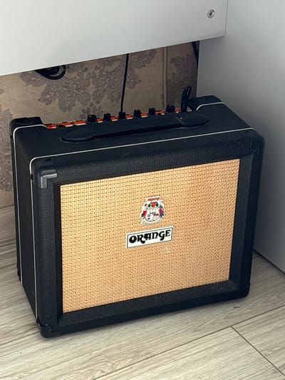 The image shows a close-up, angled view of an Orange brand guitar amplifier. The amplifier has a textured black casing with a distinctive woven tan speaker grill. A small, colorful sticker featuring cartoon characters is affixed to the speaker grill, to the left of the prominent white "ORANGE" logo, also vertically oriented. Black knobs are visible on the side of the amplifier, suggesting controls for volume or tone. The amplifier is placed on a light-colored wooden floor, with a patterned wallpaper visible behind it. To the left of the amplifier, a white surface with a silver screw is partially visible, possibly indicating a desk or shelf. The overall setting appears to be indoors, in a room with a rustic or vintage aesthetic due to the wallpaper. No people are visible in the image. The lighting suggests it is daytime, but there are no clear indicators of the time of day or weather. The context provided indicates the location as Radkevshchina, Russia.