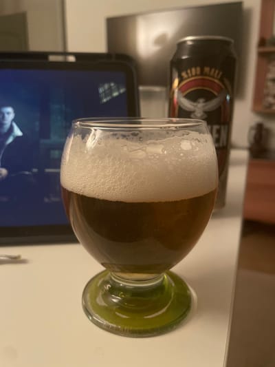 A glass of dark amber beer with a foamy head sits on a white surface, possibly a table or counter, in front of a blurred background. A black can with a gold and red label is partially visible behind the glass, suggesting it is the source of the beer. To the left, a tablet or screen displays a person's face in a dark setting. The environment appears to be indoors, likely a domestic setting or a casual establishment, with warm lighting contributing to the overall ambiance. The scene suggests a moment of relaxation or leisure. The location in Yunus Emre, Turkey, is not discernible from the visual elements of the image.