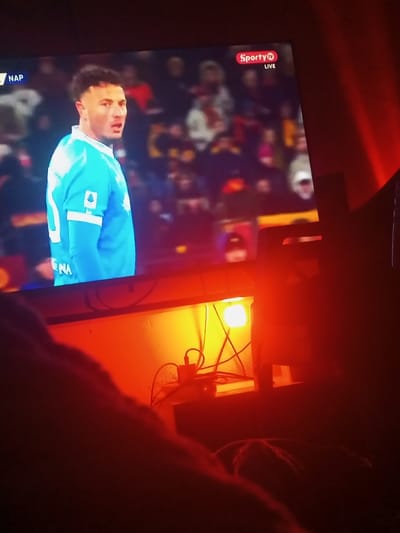 A person is watching a soccer game on a TV, which displays a player in a light blue jersey with the number 10. "Sporty TV LIVE" is visible in the top right corner. The TV is on a stand, and below it, there's a lamp casting an orange glow. Cords and devices are visible near the lamp. The room appears dimly lit, and the lower portion of the frame shows a blurred leg of the person watching TV.