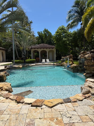This media file depicts a swimming pool area, likely in a tropical or subtropical climate given the presence of palm trees. The pool is filled with clear, turquoise water, and its edge is lined with natural stones and rock formations, giving it a resort-like feel. A small waterfall spills into the pool from one of the rock features. In the background, a covered pavilion with arched openings and a terracotta-tiled roof is visible, suggesting it's part of a larger estate or resort. Lush green trees and vegetation surround the pool area, contributing to the serene and natural setting. The sky is a clear, bright blue, indicating a sunny day. The sun is casting dappled shadows on the water and the paved area next to the pool. There are no people visible in the image, and no specific activity is taking place beyond the gentle flow of the waterfall. The overall impression is one of a tranquil and luxurious outdoor space, perfect for relaxation and recreation. The time of day appears to be late morning or early afternoon, given the bright sunlight and the shadows. The location context of Pinecrest, USA, aligns with the visual cues of palm trees and a sunny climate. There is a visible sign in blue text on a white background near the waterfall, reading "SPLASH NET 2000".