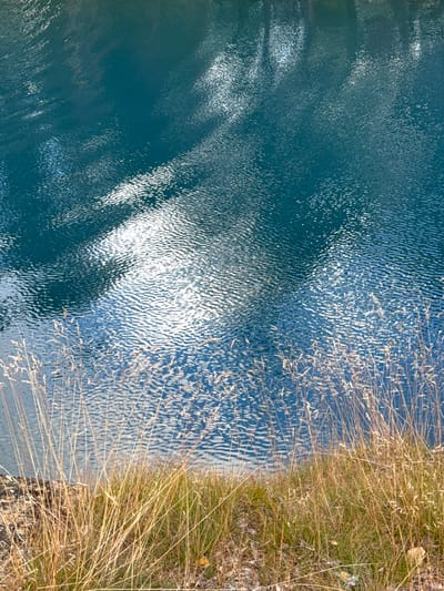The image shows a close-up view of a body of water, likely a lake or river, with a shoreline of dry grass in the foreground. The water is a deep teal color with visible ripples and reflections of the sky and surrounding environment, suggesting it is a calm day. The shoreline is covered in tall, golden-brown dry grass, indicating it might be autumn or a dry season. There are also some small rocks and fallen leaves on the ground near the grass. The scene appears to be a natural landscape. The location is not specified within the image itself, but the prompt requests it be identified as Säter, Sweden.