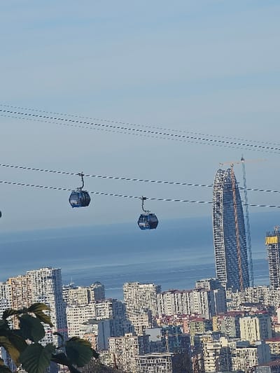 A cityscape with a partially constructed skyscraper dominates the background beside numerous other buildings. A blue sea or large body of water stretches behind the city. Two gondola cars hang suspended from wires in the foreground, against a backdrop of a clear, pale blue sky. Green foliage is partially visible in the bottom right corner of the frame.