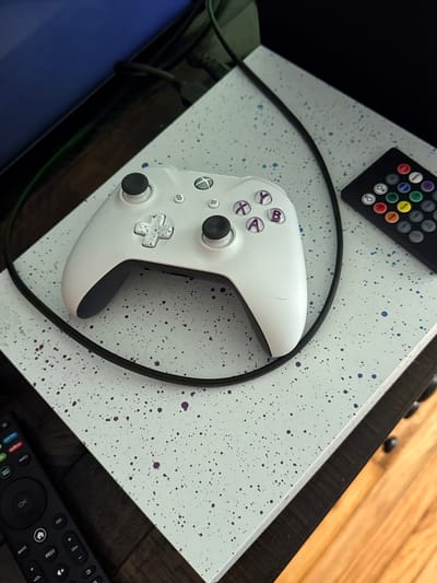 In an indoor setting, an Xbox controller and a remote control are placed on a white, speckled surface. The Xbox controller, which is white with purple buttons, sits in the center of the frame. To the right of the controller is a black remote control with numerous colored buttons. A black cord snakes around the controller and extends out of frame to the right. The surface the objects are on is light blue with dark blue and purple speckles. The background is dark and out of focus, with hints of a wooden floor visible in the lower right corner. The scene is forensic and matter-of-fact, with no people or interactions depicted. The location is not specified.
