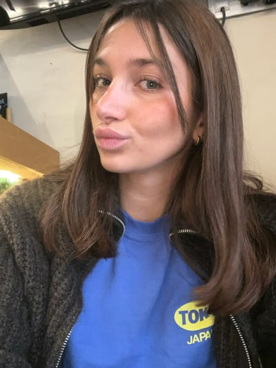 An indoor close-up selfie depicts a young adult female with long, dark brown hair, fair skin, and light-colored eyes. She is posing with puckered lips and wears small, gold-colored hoop earrings. Her attire consists of a bright blue long-sleeved shirt featuring a yellow oval logo that reads "TOKYO JAPAN," worn under a dark, textured knit zippered cardigan. The background, which is out of focus, shows elements of an interior commercial space, including a light-colored wall, a dark overhead fixture with exposed wiring, and a light wooden surface with indistinct green foliage behind it. The specific location within Aix-en-Provence, France, cannot be determined from the visual evidence.