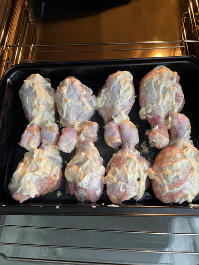The image shows a black baking tray containing eight chicken drumsticks coated in a creamy white sauce, placed inside an oven. The oven racks are visible on the left. The location Лесной Пейзаж Кп (шарапово, Russia is extraneous and irrelevant to the image content.