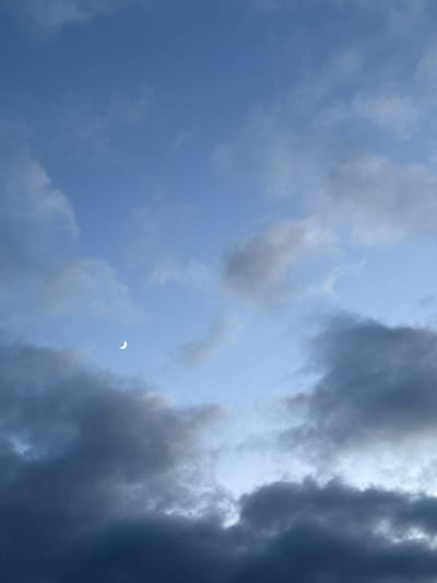 The image captures a twilight sky over Santiago de Compostela, Spain. A crescent moon is visible in the upper left quadrant of the frame, set against a backdrop of a blue sky scattered with clouds. Some clouds are dark and heavy, suggesting an approaching weather front, while others are lighter and wispy, allowing patches of blue to show through. The overall lighting indicates the transition from day to night, with the sky still holding some residual daylight. There are no people, structures, or other identifiable objects in the scene.