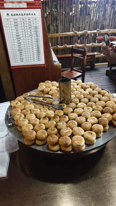 This interior image, located in Jiyang District, China, depicts a food establishment. A large, round, metallic tray containing numerous small, golden-brown baked goods occupies the center, resting on a dark wooden counter. Two metallic tongs are placed on the baked goods, alongside a central, upright cylindrical metal container. To the left, a white sign with Chinese text, identified as "Huayuan Convenience Store Product Price List," is mounted on a wooden display stand. The background features a wall constructed of vertical bamboo poles and horizontal wooden rails. A wooden chair and a small wooden table are partially visible behind the food display. On the right, a person's hand, partially visible in a patterned sleeve, holds a red-handled tool near the bamboo wall. Handwritten notes on paper are situated on the counter to the immediate left of the food tray.
