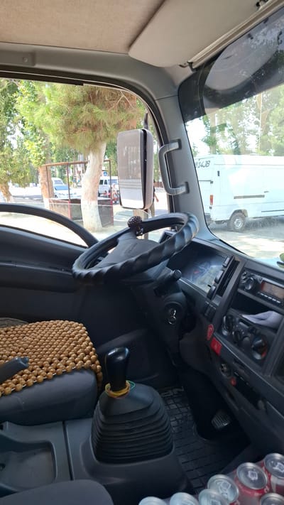 The media file shows the interior of a truck cabin, viewed from the passenger's side, looking towards the driver's side and out the windshield. The primary subjects are the truck's interior components and the scene outside.

Inside the cabin, the steering wheel, dashboard with various controls and a radio, and the gear stick are clearly visible. A beaded seat cover is draped over the driver's seat. A rearview mirror is mounted on the windshield, reflecting the outside scene. Several cans of soda are stacked on the right side of the cabin, near the dashboard.

Through the windshield and side windows, a street scene is visible. Trees with green foliage are prominent, suggesting it is daytime and likely spring or summer. There are other vehicles, including what appears to be a white bus or truck, parked on the street. The reflection in the rearview mirror also shows similar outdoor elements.

There is some visible text on the steering wheel, which appears to say "ISUZU". On the exterior of the white vehicle visible through the windshield, there is text that reads "914JB".

The scene appears to be a snapshot of a typical day in a vehicle, possibly parked on a street or lot. No specific activity or event is depicted, beyond the general context of being inside a vehicle. The lighting suggests it is daytime, and the overall impression is casual and ordinary. The location context of Kuvasay, Uzbekistan, indicates the geographical setting of the image.