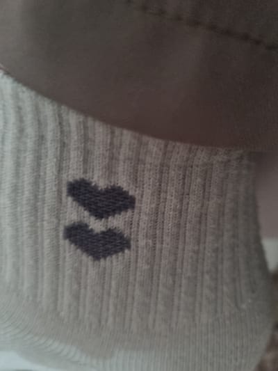 The image shows a close-up of a light grey sock with a ribbed texture. Two dark grey chevron shapes are embroidered on the sock, one above the other, forming a stylized arrow or two connected arrows. The sock appears to be made of a soft, knitted material. The background is blurred and out of focus, showing a neutral, possibly skin-toned or fabric surface. There is no identifiable information within the image to suggest a specific location in Shopokov, Kyrgyzstan, or any action or interaction.