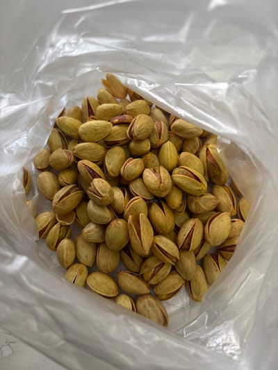 A clear, crinkled plastic bag is depicted from an elevated perspective, filled with numerous in-shell pistachios. The pistachios feature light tan to yellowish-green shells, with many naturally split open to reveal reddish-purple and green kernels. The interior of the bag is predominantly filled with these nuts, with slight variations in shell color and kernel visibility. The scene is located in Lehvaz, Armenia.