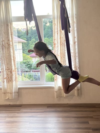 The image shows a young girl performing aerial yoga in a room with large windows. She is suspended in the air, supported by a dark purple aerial silk, with her legs extended and one foot pointed upwards. She is wearing a light blue t-shirt and grey shorts, and yellow socks. Her hair is dark and pulled back.
The room has floor-to-ceiling windows that look out onto a lush green outdoor scene, with trees and glimpses of houses. The light suggests it is daytime, possibly late afternoon given the warm glow. The curtains are white with a floral pattern. The floor is made of wood planks.
The girl appears focused on her movements, her body contorted into an athletic pose. There are no visible signs of text. The overall impression is one of active movement and peaceful focus in a bright, natural setting. The location is Verkhniy Yurt, Russia.