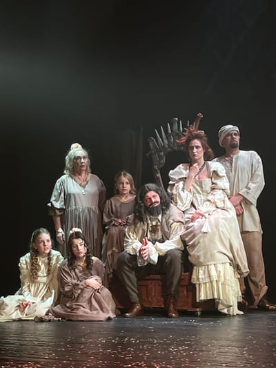 This image captures seven individuals posed on a dimly lit stage, consistent with a theatrical performance at a venue in Novoshakhtinsk, Russia. The background is predominantly dark, with a skeletal-like, dark-colored set piece visible behind the central figures. The stage floor is dark, scattered with light-colored debris.
In the foreground, two young females are seated on the floor. On the left, a girl with light hair in a light-colored, long-sleeved dress. Next to her, a young woman with dark hair wears a dark brown, long-sleeved dress, seated with her knees drawn up.
Behind them, an older woman with white hair pulled back stands, wearing a long, light grey dress with a pendant, her mouth slightly open and eyes wide. To her immediate right, a young girl with blonde hair stands, wearing a plain dark brown dress.
Centered on a dark wooden chest or bench, a man with long dark hair and a full dark beard is seated. He wears a light-colored, ruffled shirt, a darker vest, dark trousers, and brown boots, with his hands clasped. To his right, a woman is also seated on the chest. She has dark hair styled in an elaborate updo with a decorative element and a small, pointed crown-like accessory. She wears a voluminous light-colored gown with ruffled sleeves and a dark necklace, with her left hand near her chin.
Standing to the far right, behind the seated woman, is a man wearing a light-colored turban, loose tunic, and trousers. All subjects are looking generally forward.