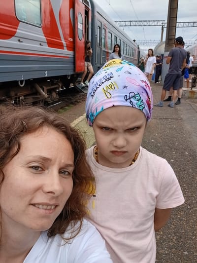The image captures a selfie of two people, an adult woman and a young girl, with a train and a train station platform in the background. The woman, on the left, has curly brown hair and is smiling. The young girl, on the right, is wearing a colorful headscarf and has an angry expression. She is wearing a pink t-shirt. In the background, people are seen standing on a train platform next to a red and gray train. The environment appears to be outdoors, with a cloudy sky overhead. The location is identified as the Kropachyovo, Russia train station.
