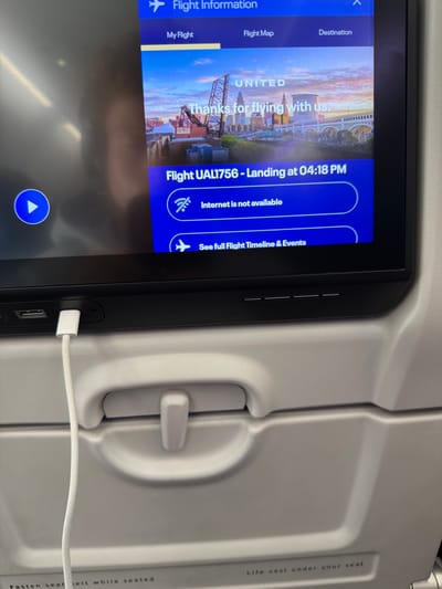 An eye-level shot captures a gray airplane seat and a black seat-back entertainment screen within an airplane cabin. The screen displays flight information for "UNITED" flight "UAL1756", showing "Landing at 04:18 PM" and a message indicating "Internet is not available." The background image on the screen is a vibrant sunset cityscape, resembling Cleveland with its industrial waterfront and prominent buildings, alongside the welcoming text "Thanks for flying with us." Navigational tabs such as "Flight Information," "My Flight," "Flight Map," and "Destination" are visible on the left.

A white braided charging cable is plugged into a USB port located on the lower right side of the screen's frame, suggesting a device is being charged. The textured gray plastic of the airplane seat is visible, including a small latch or handle for the tray table. Text printed on the side panel of the seat reads "Life vest under your seat" and "Fasten seat belt while seated." The scene depicts a passenger in transit during an afternoon flight, utilizing the in-flight entertainment system.