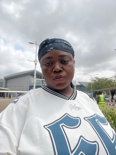 This image is a low-angle selfie, rotated 90 degrees clockwise, featuring a person with dark skin. The subject looks directly at the camera with a neutral expression. They are wearing a dark, patterned headscarf and a white short-sleeved top, possibly a sports jersey, with black and white striped trim. The shirt prominently displays large blue numbers, likely "23," outlined in black, and a smaller cursive text reading "Kings" on the upper left chest area. A small gold hoop nose piercing is visible.
The background shows an outdoor setting under a cloudy, overcast sky, indicating daytime. Modern, multi-story buildings with grey and glass facades are visible, suggesting a large urban complex or an airport terminal. Green trees, bushes, and several utility poles or streetlights are also present. In the mid-ground, another person wearing a bright yellow/lime green high-visibility vest can be seen, likely a worker or security personnel. The scene suggests a public space, consistent with an airport or large facility in Gwaska, Nigeria.