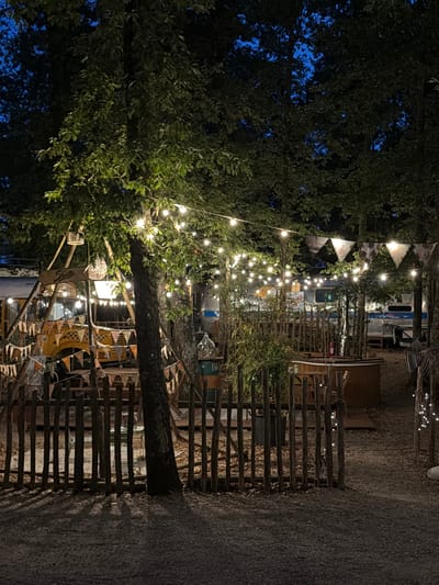 The image depicts an outdoor scene at night, illuminated by strings of fairy lights and the faint glow of a blue sky peeking through the trees. The environment appears to be a campsite or outdoor venue nestled amongst trees. In the foreground, a rustic wooden fence made of vertical posts and horizontal rails encloses an area with gravel and fallen leaves. Beyond the fence, a yellow vehicle, possibly a converted school bus or truck, is partially visible, adorned with colorful triangular flags. Several other structures, including what looks like a caravan or RV, are present in the background. The overall atmosphere is dimly lit and cozy, suggesting a relaxed evening setting. The location is identified as Castelnuovo dei Sabbioni, Italy.