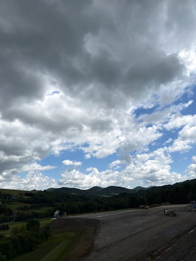 The image depicts a construction site located in the rural area of Stróża, Poland. The foreground is dominated by a large, unfinished construction area with dark, exposed earth and gravel. Several construction vehicles, including a large yellow dump truck and a green tractor with a trailer, are present on the site. In the mid-ground, there are rolling green hills covered with lush trees. Scattered among the hills are a few houses, indicating a nearby residential area. The background features a range of distant, tree-covered mountains under a dramatic sky filled with a mix of dark, heavy clouds and patches of bright blue sky with fluffy white clouds. The overall scene suggests a large-scale construction project taking place in a scenic, natural environment.