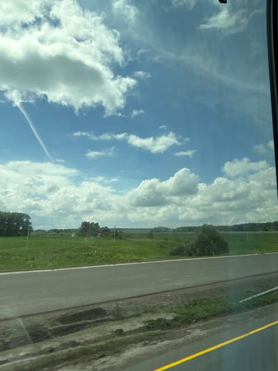 The image depicts a rural landscape viewed from inside a vehicle, likely a car or bus, given the perspective through a window. The scene showcases a vast expanse of green fields under a bright blue sky dotted with fluffy white clouds and a thin contrail from an aircraft. A paved road with a yellow line runs horizontally across the foreground. The background features scattered trees and vegetation, suggesting a natural, undeveloped area. The location is identified as Taimurazino, Russia. There are no people or animals visible in the image. The overall impression is one of a serene, open, and natural environment on a clear day.