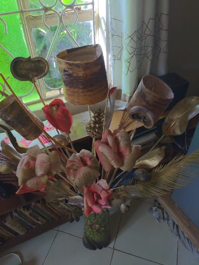 A bouquet of artificial flowers, consisting of gold and red silk blossoms, is arranged in a green and gold ceramic vase on a shelf next to a window. The flowers are arranged in a vase on a shelf. The vase is made of ceramic and is painted green and gold. A white curtain with a gold geometric design hangs over the window, and a portion of a wooden bookcase is visible in the background, with several old books on the shelf. The bouquet is positioned next to the window. The window is open and the view outside is of a tropical jungle. The image is taken from above. This image appears to have been taken in a home in Lindi Street, Tanzania, near the coordinates -3.357 latitude, 37.333 longitude.