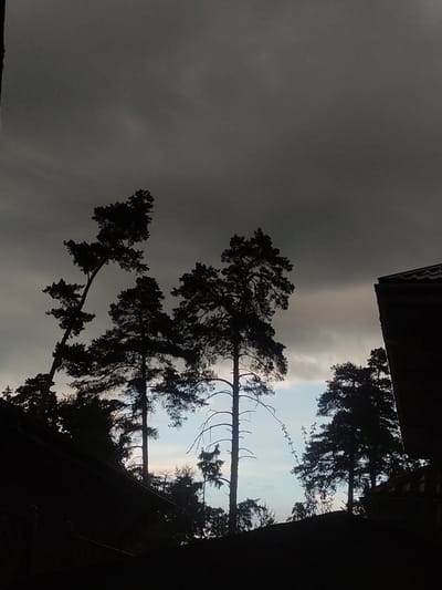 The image shows a view upwards towards a sky with heavy, dark clouds. Patches of lighter sky are visible amidst the dark clouds, hinting at a possible clearing. Silhouetted trees are in the foreground, framing the sky and the buildings on top of the image. The angle of the shot suggests it was taken from ground level. The general impression is dark and overcast. The location in the city of Kratovo, Russia is not determinable from the visual information provided.