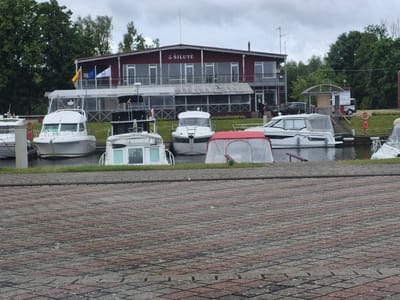 The image depicts a waterfront scene in Šilutė, Lithuania, likely at a marina or harbor. In the foreground, a paved area with diamond-patterned concrete blocks forms the ground. Beyond this, a body of water, likely a river or bay, is visible, lined with green grass. Moored boats of various sizes are present on the water.
Prominently featured is a two-story building with a red facade and gray trim, labeled "ŠILUTĖ" in white lettering. This building appears to be a restaurant, club, or public facility, with large windows and balconies. Flags, including the Lithuanian flag and the European Union flag, are displayed on poles in front of the building.
Several boats are docked. On the left, a white motor yacht is moored. Next to it, another white boat with a covered cabin is visible. A white boat with a red canopy is in the center, and to its right, a larger white motor yacht with the name "Maya" on its side is docked. Further to the right, another white boat with a canopy is partially visible. A dark-colored car is parked near the building on the right side of the image.
The environment is overcast, suggesting a cloudy day. Trees and vegetation are visible behind the building and on the surrounding land. The scene conveys a sense of a recreational area by the water.