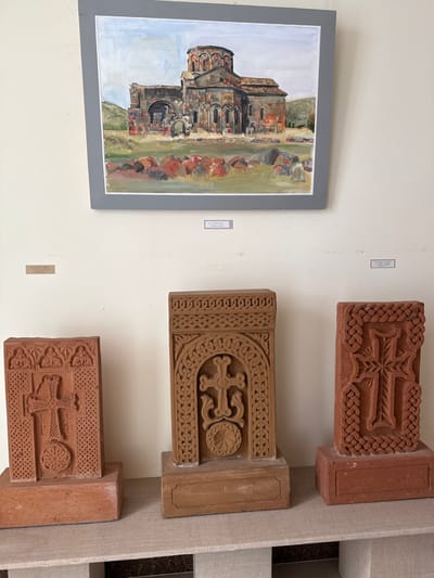 An indoor scene displays cultural artifacts against a plain light-colored wall, likely within a museum or exhibition space in Talin, Armenia. Centrally positioned on the wall is a framed painting, depicting the ancient, partially ruined Talin Cathedral, an edifice of dark stone with a central dome, set amidst a sparse landscape with scattered reddish and gray rocks under a light blue sky. Below this painting, three carved stone monuments, identified as khachkars (Armenian cross-stones), are exhibited on a light-colored plinth. The leftmost and rightmost khachkars are rectangular and reddish-brown, each featuring a prominent ornate cross with a circular element at its base, encased within decorative borders. The central khachkar is taller and lighter in tone, displaying an elaborate cross with swirling motifs and a circular base, enclosed within an intricately carved arched frame with braided patterns. Small white labels with black text are affixed to the wall below the painting, two of which partially read "TALIN...", confirming the geographical context. No individuals are present in the scene.