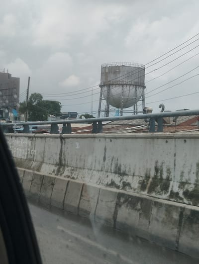 The image depicts an outdoor scene on a cloudy day, likely in Alagbede, Nigeria. The main subject is a large, cylindrical water tank supported by a metal structure, positioned prominently in the mid-ground. The tank appears weathered, with visible signs of age and possibly rust.

In the foreground, a concrete barrier or wall runs horizontally across the frame, showing signs of wear and tear, with patches of moss or algae. On this wall, some text is visible, appearing to be spray-painted or written in chalk. The legible parts read "CLEANER-40K" and a phone number that seems to start with "070...".

Behind the water tank, a cluster of trees with dense green foliage can be seen. Further in the background, other structures, possibly buildings and antennas, are visible, along with a network of overhead power lines that crisscross the sky. The overall impression is of an urban or semi-urban environment with industrial elements.

The weather appears overcast, indicated by the diffused light and the grey sky. The mood is neutral, with no distinct emotional cues. The time of day is not clearly identifiable due to the cloud cover. The presence of the water tank suggests a utility or industrial area. The low angle of the shot, possibly taken from a moving vehicle, gives a sense of perspective and the busyness of daily life in the location.