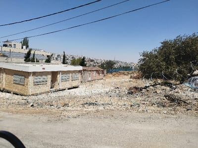 The image captures an outdoor scene under a clear, bright blue sky in Al-Khalil, Palestinian Territory.

The foreground consists of a light-colored dirt and gravel road, partially obscured by a dark, rounded object on the lower left, possibly part of a vehicle. Immediately beyond the road, the terrain is unpaved, arid, and appears littered with various debris and multiple coils of concertina wire that extend horizontally across the lower half of the frame.

In the midground, to the left, a single-story, light-colored modular building with a flat roof is present. Its walls appear to be constructed from light-colored panels. Three windows on the visible side are fitted with closed, horizontal slatted shutters. A red rectangular sign is affixed above a central entrance door. Behind this structure, a smaller, older-looking, reddish-brown building with a sloped roof is partially visible. To the right of these buildings, the unpaved, debris-strewn area continues, leading to a dense cluster of dark green trees and bushes that occupy the right side of the frame. Additional concertina wire is present near the base of these trees.

The background, from left to center, reveals a hilltop settlement featuring several multi-story, light-colored buildings, some equipped with arrays of solar panels on their roofs. Narrow, upright green trees are interspersed among these structures. Further right, a sprawling urban landscape of densely packed, light-colored buildings stretches across distant hills under the bright sunlight. A blue barrier or fence is discernible between the midground and the distant city.

No individuals or active movements are observed within the scene.