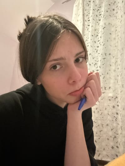 A close-up, eye-level indoor portrait features a young individual with dark brown, medium-length hair and fair skin. The image is oriented with the person's head tilted 90 degrees to the right. The person is looking directly into the camera with wide, engaging eyes. They are wearing a plain black long-sleeved shirt or sweater.
Their right hand is raised, with fingers curled, supporting their chin or jaw. A bright blue pen is held between their lower lip and chin, with the tip resting against the mouth area, suggesting a pensive or thoughtful posture, or perhaps boredom. The lips are slightly parted around the pen.
In the background, a white or light-colored curtain or fabric with a delicate, repetitive floral or geometric pattern is visible, illuminated by soft, diffused light. The setting appears to be a domestic indoor space, with no specific time of day discernible beyond indoor lighting. There is no visible text in the image. This scene is set in Volkhovets, Russia.
