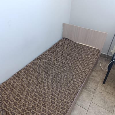 The image displays an interior room located in the city of Hlybokaye, Belarus. A single bed with a light wood-grain headboard and frame is centrally positioned, abutting a plain, light-colored wall. The mattress is covered by a textile featuring a repetitive geometric pattern in brown and beige, creating an optical illusion of interconnected cubes. The floor is composed of square, light-toned tiles. To the right of the bed, a dark-colored chair leg is visible, along with a white electrical cord extending towards the wall. A vertical metallic pipe is affixed to the wall in this area. No individuals, actions, or interactions are present in the scene.