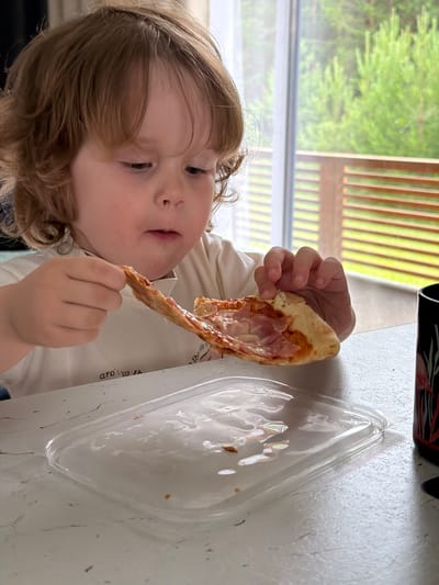 In Kamyennoye, Russia, a young child with curly brown hair sits at a table, holding a slice of pizza. The child, wearing a cream-colored t-shirt with text on it, is looking down at the pizza with a focused expression, a piece of cheese hanging from the crust. In front of the child is a clear plastic container with some remnants of food, and to the right, a black cup with a floral pattern is visible. The background shows a large window with a view of green trees and a wooden railing, suggesting an indoor setting with an outdoor view. The table surface appears to be a light-colored material with visible texture and some dark specks.